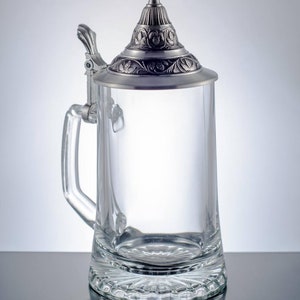 May include: A clear glass beer stein featuring a heavy base with a radial pattern and a hinged pewter lid. The lid is conical with an embossed floral and leaf design, complemented by a matching metal thumb lever attached to the glass handle.