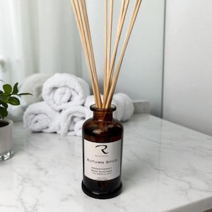 May include: A brown glass aromatherapy reed diffuser with wooden reeds. The label reads "Autumn Spice" and "Aromatherapy Reed Diffuser with Essential Oils". The diffuser is on a marble surface, with rolled white towels in the background.