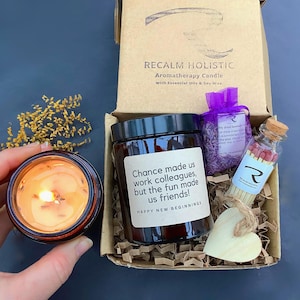 May include: Aromatherapy candle gift set in a cardboard box. The set includes a lit candle in a glass jar, a jar of matches, a small bag of dried lavender, and a wooden heart. The candle jar has the text "Chance made us work colleagues, but the fun made us friends!"