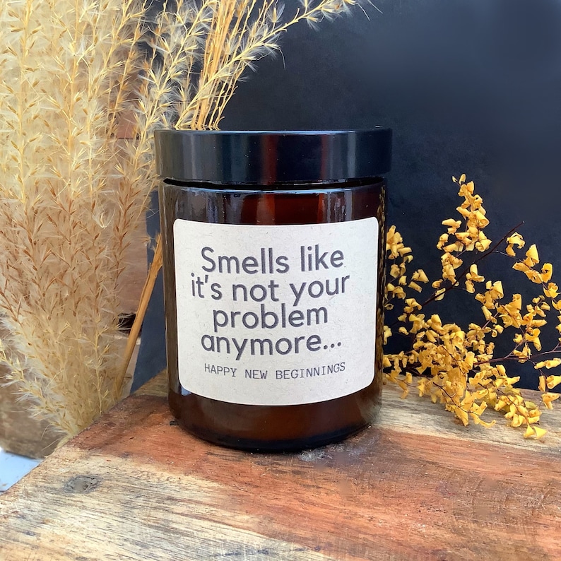Pode incluir: Uma vela de vidro marrom com um r&oacute;tulo branco que diz "Smells like it's not your problem anymore... Happy New Beginnings".
