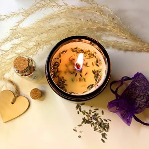 May include: A lit candle in a brown glass jar with a white wax base. The candle has dried lavender flowers and herbs embedded in the wax. The candle is surrounded by dried lavender flowers.