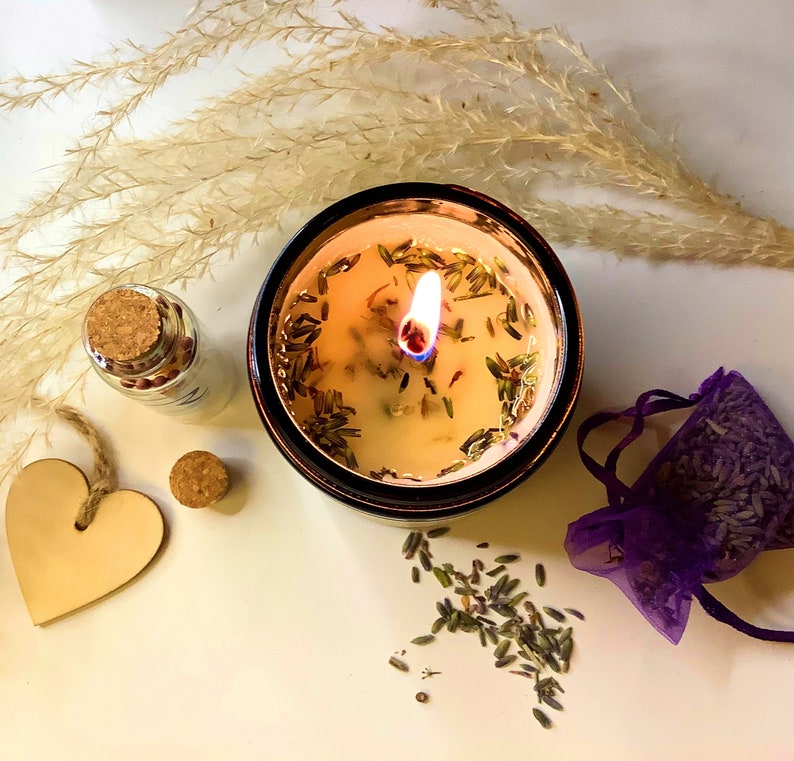 May include: A lit candle in a brown glass jar with a white wax base. The candle has dried lavender flowers and herbs embedded in the wax. The candle is surrounded by dried lavender flowers.