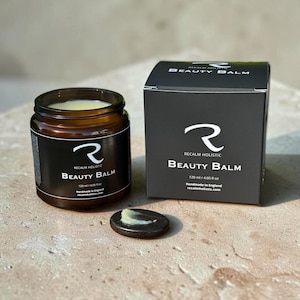 Pode incluir: Um frasco de vidro castanho e uma caixa de cartão preta de bálsamo de beleza Recalm Holistic, ambos rotulados com 120 ml / 4,05 fl oz. Uma pequena pedra escura e lisa está à frente do frasco com um pouco de bálsamo amarelo pálido.