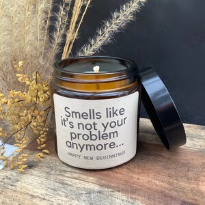 Pode incluir: Uma vela de vidro marrom com um r&oacute;tulo branco que diz "Smells like it's not your problem anymore... Happy New Beginnings". A vela est&aacute; sentada em uma superf&iacute;cie de madeira com flores secas no fundo.