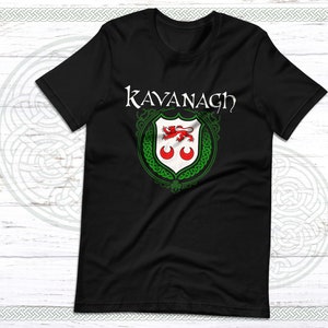 May include: Black t-shirt featuring the name "Kavanagh" in white script above a family crest. The crest includes a red lion, two red crescents, and green detailing. The shirt is displayed on a white wooden surface.