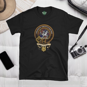 Puede incluir: Camiseta negra con un escudo de clan escocés dorado y rojo con un dragón y el texto "Conjuctu Virtuti Fortuna" y "Macbeth".