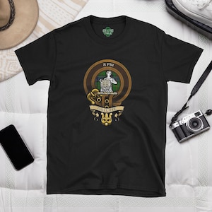 Peut inclure: T-shirt noir avec un blason de clan or et vert représentant une femme tenant un bouclier. Le blason est entouré d'un cercle doré avec le texte "OGILVIE" et "A FIN" en lettres dorées.