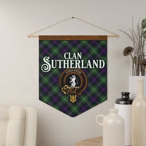 May include: A wall hanging banner displaying the words "Clan Sutherland Sans Peur" in white. The banner has a green, blue, and red tartan pattern. A crest with a white lion is centered below the text. It is hung from a wooden rod.