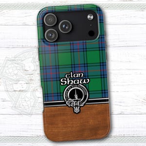 May include: A phone case featuring a green, blue, and red plaid pattern with the text "clan Shaw" and a crest. The bottom portion has a wood grain design. The case has a black camera module.