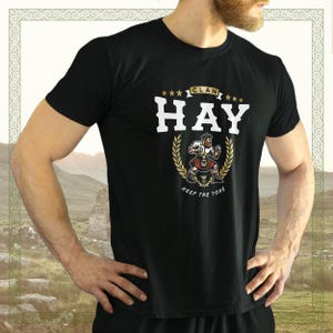 Op de afbeelding: Zwart T-shirt met een witte afbeelding van een man in een kilt met de tekst "Clan Hay Keep the Yore" in gouden letters.