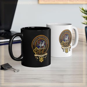 Puede incluir: Dos tazas de cerámica, una negra y otra blanca, cada una con un escudo heráldico. El escudo presenta un dragón, una pancarta con la palabra "Macbeth" y el lema "Conjuncta Virtuti Fortuna". Las tazas están sobre una superficie clara.