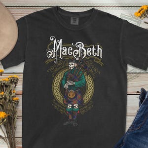 Puede incluir: Camiseta negra con un diseño de un esqueleto tocando la gaita, vestido con una chaqueta verde y un kilt a cuadros. La palabra "MacBeth" está escrita encima de la ilustración. El diseño está dentro de un círculo de nudos celtas dorados.