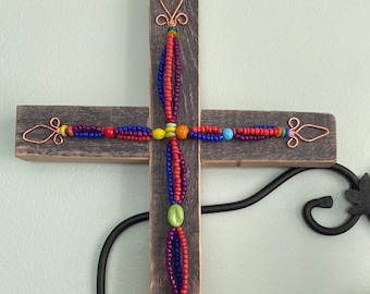 May include: A wooden cross with a beaded design. The beads are red, blue, green, and yellow. The cross is decorated with copper wire.