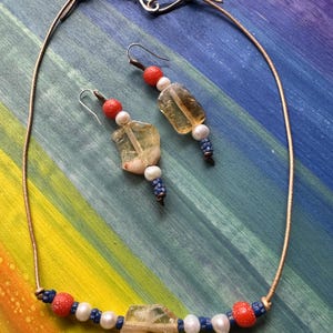 May include: A leather necklace with a large, clear, yellow gemstone pendant. The necklace is accented with red and white beads. Matching earrings with a smaller version of the pendant and red and white beads.