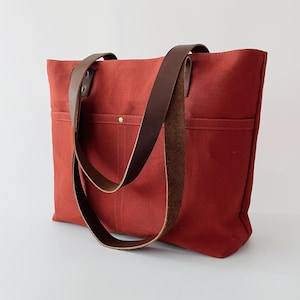 May include: A red canvas tote bag with brown leather straps. The bag has two exterior pockets and a brass rivet detail. The leather straps are attached to the bag with brass rivets. The bag is designed for carrying items.
