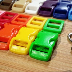 May include: Assortment of colorful plastic side release buckles in various sizes. The buckles are arranged in a row on a wooden surface. There are also several silver metal D-rings.