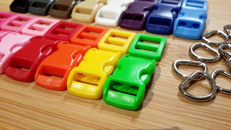 May include: Assortment of colorful plastic side release buckles in various sizes. The buckles are arranged in a row on a wooden surface. There are also several silver metal D-rings.