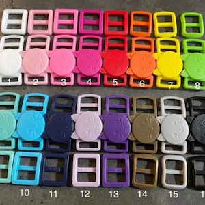 May include: A set of 16 plastic slider buckles with a cat face design. The buckles are in various colors, including white, pink, red, orange, yellow, green, blue, purple, brown, and black. The cat faces are all smiling and have a small nose and whiskers.