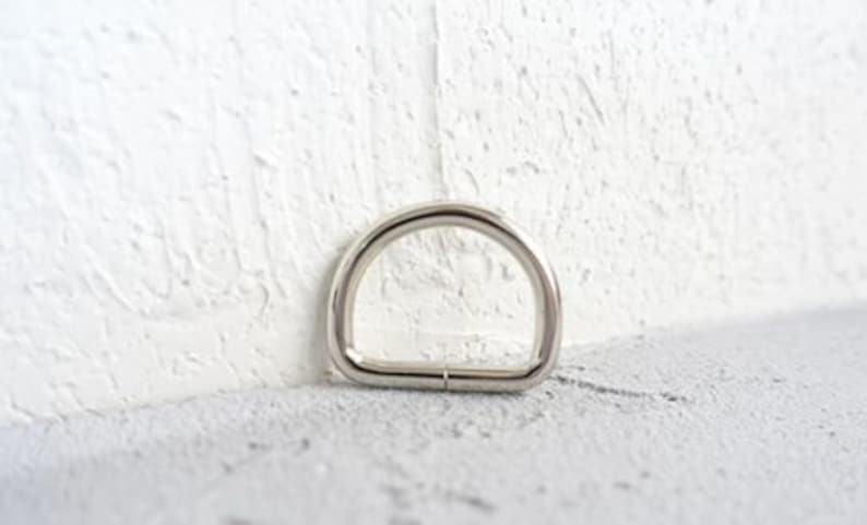 Op de afbeelding: Een zilveren metalen D-ring, vaak gebruikt om banden of handvatten aan tassen, riemen of andere accessoires te bevestigen.