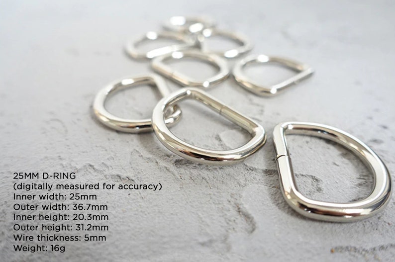 Op de afbeelding: Een close-up van tien zilveren 25 mm D-ringen. De ringen zijn gemaakt van metaal en hebben een gladde, gepolijste afwerking. De tekst "25MM D-RING (digitally measured for accuracy) Inner width: 25mm Outer width: 36.7mm Inner height: 20.3mm Outer height: 31.2mm Wire thickness: 5mm Weight: 16g" is zichtbaar onder de ringen.