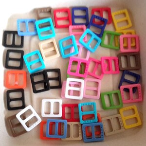 May include: Assortment of colorful plastic slider buckles in various colors, including red, blue, green, yellow, black, white, and brown. The buckles are all the same size and shape, and they have a rectangular design with two holes in the center.