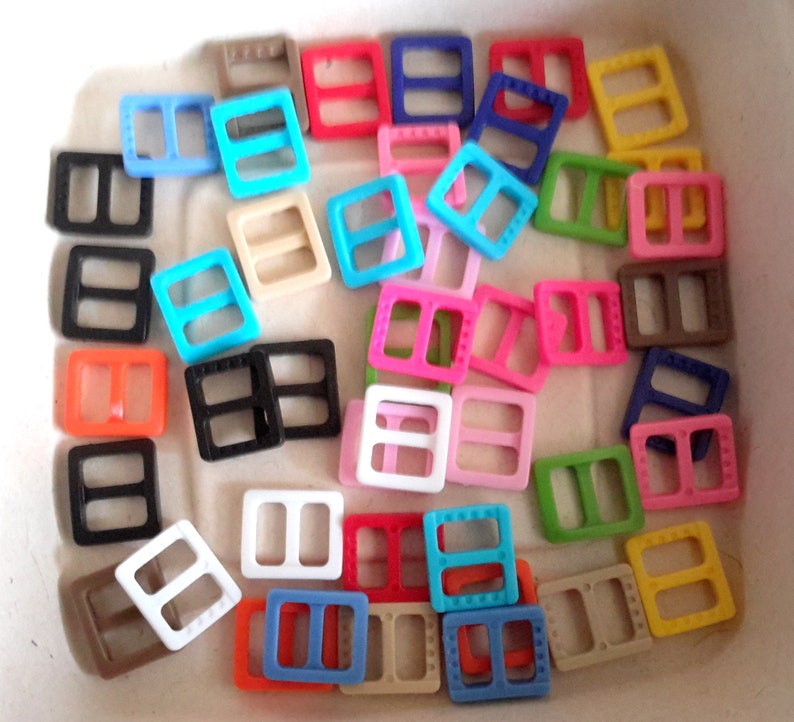 May include: Assortment of colorful plastic slider buckles in various colors, including red, blue, green, yellow, black, white, and brown. The buckles are all the same size and shape, and they have a rectangular design with two holes in the center.
