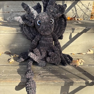 May include: A plush, dark gray axolotl stuffed animal with large, round eyes and feathery gills. The handmade creature has a long, segmented tail and is posed on wooden steps. The soft toy is a unique, whimsical creation.