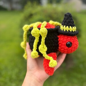 Puede incluir: Un juguete de araña de ganchillo hecho a mano en negro, rojo y amarillo. La araña tiene un sombrero de bruja y sostiene un caldero. El cuerpo de la araña es negro, con una cabeza y patas rojas. Las patas de la araña son amarillas.
