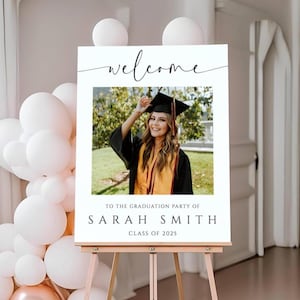 May include: A graduation party welcome sign on a wooden easel. The sign features a photo of a smiling graduate in a cap and gown, with the word "welcome" in script above the photo. The text below reads "To the graduation party of Sarah Smith, Class of 2025."