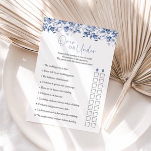 May include: A printable wedding shower game card with a blue floral design. The card has the title "Over or Under" and asks guests to guess if the number in each question is over or under the given number. The card includes 10 questions about the wedding.
