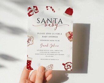 May include: A baby shower invitation with a festive Santa theme. The card features the words "Santa Baby" in elegant script, along with illustrations of Christmas ornaments. The invitation is held by a person.