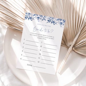 May include: A printable bridal shower game card with a blue floral design. The card has the text "How old was the Bride to be?" and asks guests to guess the bride's age in 12 photos. The person with the most correct answers wins.