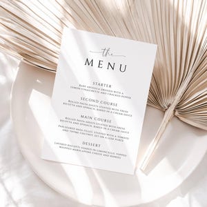 May include: A white menu card with the text "the MENU" in elegant script. The menu lists a starter, second course, main course, and dessert. The card is on a white plate with a dried palm leaf in the background.