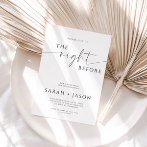 May include: A white invitation card with black text that reads "The Night Before" and details for a rehearsal dinner. The card is on a white plate with a dried palm leaf in the background.