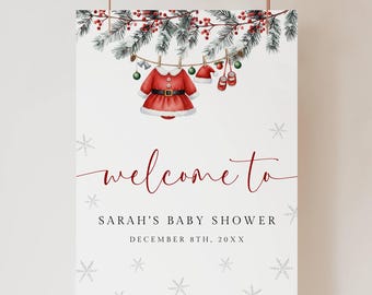 May include: A white sign with a Christmas theme, featuring a red Santa-style dress, hat, and shoes hanging from a garland. The sign reads "welcome to" in cursive, followed by "Sarah's Baby Shower" and the date.
