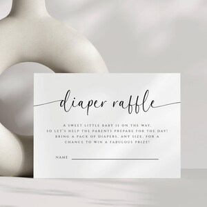May include: A white baby shower diaper raffle card with elegant script text. The card reads "diaper raffle" and includes text inviting guests to bring diapers for a chance to win a prize. A decorative white vase is in the background.