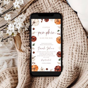 May include: A smartphone displays a baby shower invitation with a pumpkin theme. The invitation features the text "A little pumpkin is on the way" with watercolor pumpkins and foliage. The phone rests on a cozy, cream-colored knitted blanket.