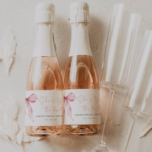 May include: Two mini bottles of sparkling wine with pink ribbon and watercolor bow accents. The labels read "She's tying the knot" and "Sarah's Bridal Shower".