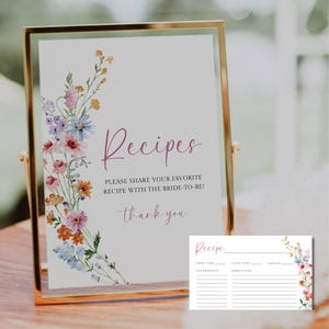 Könnte beinhalten: Ein gerahmtes Schild mit dem Wort "Rezepte" in Schreibschrift, zusammen mit dem Text "TEILEN SIE IHR LIEBLINGSREZEPT MIT DER BRAUT!" und "Danke." Das Schild ist mit einem floralen Design verziert. Eine Rezeptkarte ist ebenfalls enthalten.
