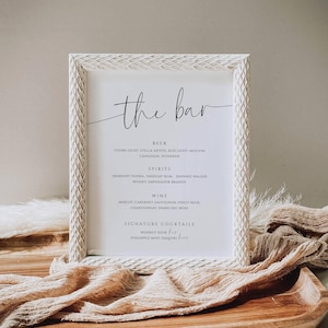 Könnte beinhalten: Ein weiß gerahmtes Schild mit dem Text "the bar" in eleganter Schrift. Das Schild listet Bier, Spirituosen, Wein und Cocktails auf. Der Rahmen hat ein strukturiertes, geschnitztes Design. Das Schild wird auf einem Holztablett mit Stoff präsentiert.