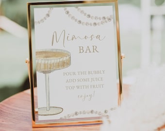 May include: A gold-framed sign with a white background and a watercolor illustration of a mimosa glass. The sign reads "Mimosa Bar" and includes instructions for making a mimosa: "Pour the bubbly, add some juice, top with fruit, enjoy!"