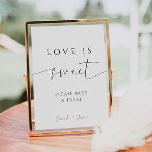 May include: A gold-framed sign with the words "LOVE IS sweet" in elegant black script. Below, it reads "PLEASE TAKE A TREAT" and the names "Sarah + Jason". The sign is displayed on a wooden surface.