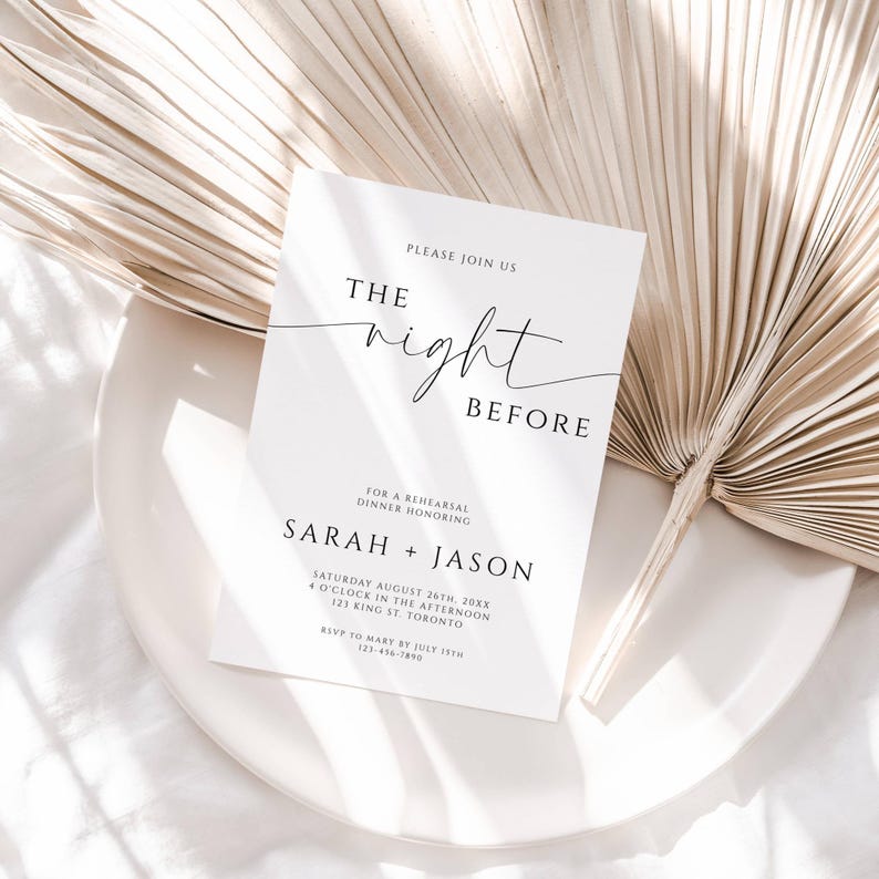 May include: A white invitation card with black text that reads "The Night Before" and details for a rehearsal dinner. The card is on a white plate with a dried palm leaf in the background.