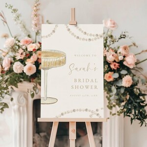 Może przedstawiać: Biały znak z ilustracją kieliszka do szampana i tekstem "Witamy na Bridal Shower Sarah 14 sierpnia 2023". Znak jest ozdobiony girlandą pereł i stoi na drewnianym sztalugach.