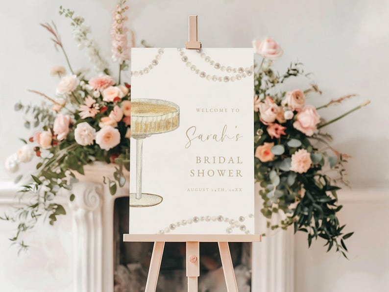 Może przedstawiać: Biały znak z ilustracją kieliszka do szampana i tekstem "Witamy na Bridal Shower Sarah 14 sierpnia 2023". Znak jest ozdobiony girlandą pereł i stoi na drewnianym sztalugach.