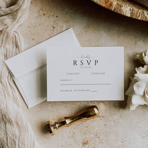 May include: A flat lay featuring a white RSVP card with the text "RSVP" and a gold wax seal. An envelope and a piece of white fabric are also present. The card includes fields for accepting or declining and the number attending.