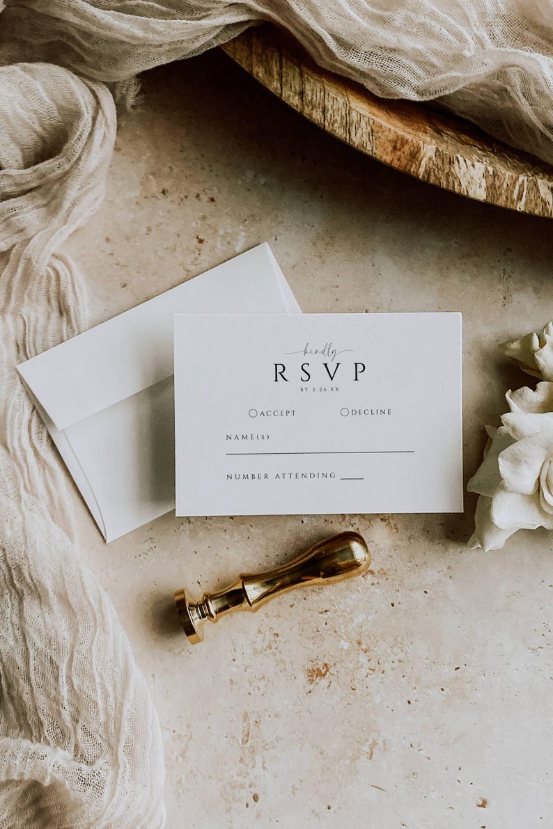 May include: A flat lay featuring a white RSVP card with the text "RSVP" and a gold wax seal. An envelope and a piece of white fabric are also present. The card includes fields for accepting or declining and the number attending.