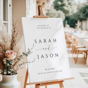 May include: White wedding welcome sign with black text. The sign reads "Welcome to the wedding of Sarah and Jason 08.26.20XX We're so glad you're here."