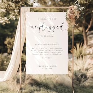 May include: A wedding ceremony sign with the words "Welcome to our unplugged ceremony." The sign is on a gold metal stand, decorated with flowers and draped fabric. The sign asks guests to turn off phones and cameras.