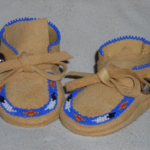 May include: A pair of tan suede baby moccasins with blue beaded trim. The moccasins have a leather tie and a small silver bell attached. The toe area features a beaded design in blue, white, black, and orange.